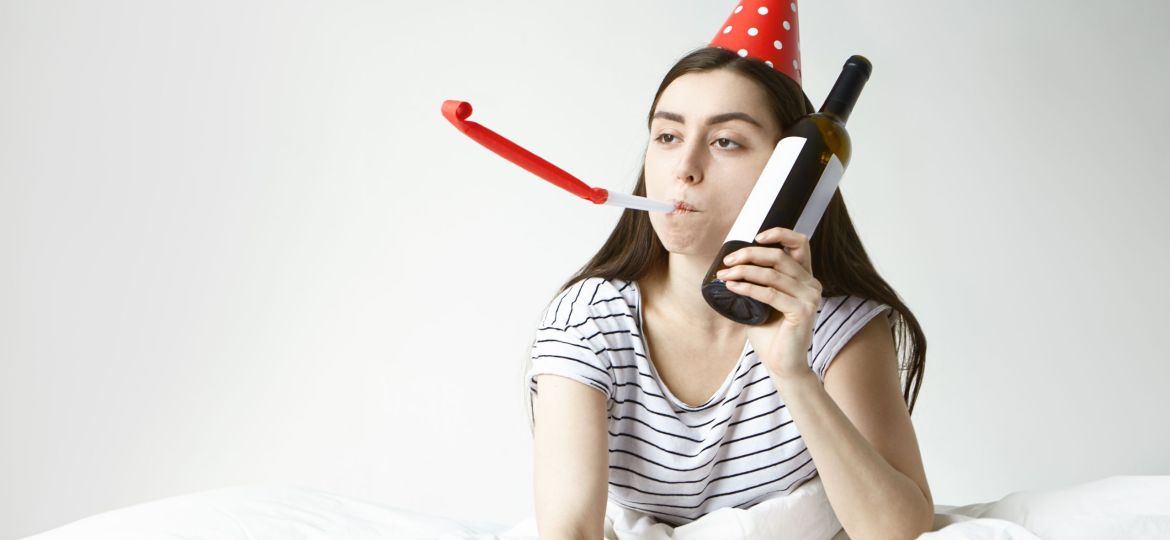 Booze, alcohol and sobriety concept. Drunken young Caucasian woman in striped pajamas awakened early in the morning after party, suffering from hangover, drinking wine from glass bottle to get better